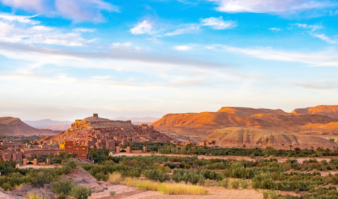 Uma vista panorâmica de Ait Benhaddou, uma aldeia histórica de barro e terra, em contraste com as colinas áridas sob um céu de pôr do sol em tons pastéis.